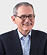 A senior person wearing round glasses, a dark blazer, and a striped shirt is smiling and clasping their hands. Description generated by AI