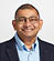 A person wearing glasses, a light blue button-up shirt, and a dark blazer is smiling with hands in pockets against a plain light background. Description generated by AI