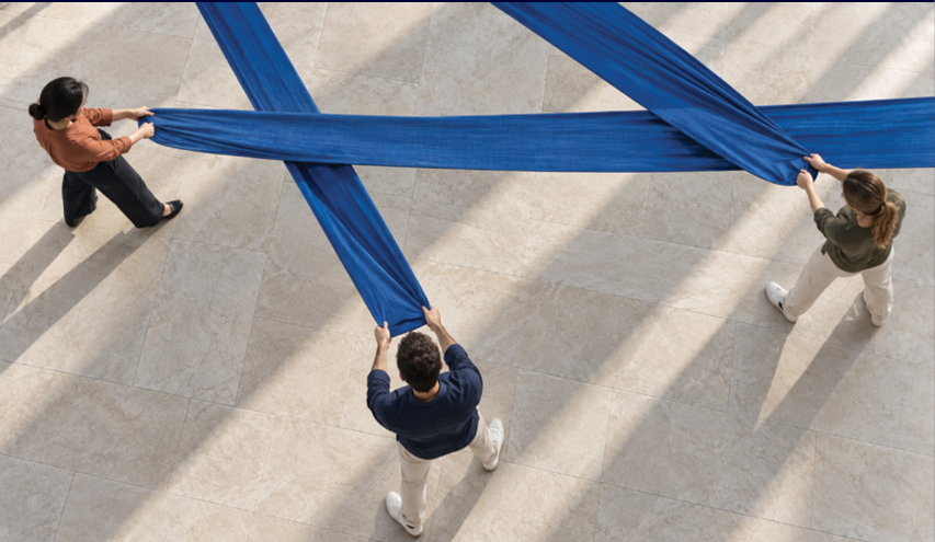 Three adults stand on a light-colored floor holding and stretching long blue fabric strips in a crisscross pattern. Description generated by AI