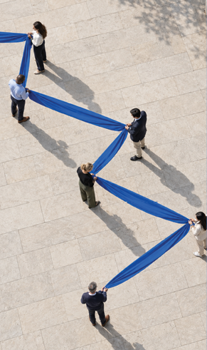 Seven people stand on a large tiled surface holding a long blue fabric stretched in a zigzag pattern. Description generated by AI