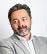 A person with short dark hair and a beard wearing a light gray blazer and white shirt is smiling gently against a plain white background. Description generated by AI