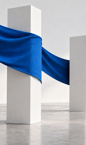 A flowing blue fabric is wrapped around a tall white rectangular pillar standing on a smooth gray floor against a plain light gray wall. Description generated by AI